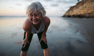 Woman with short gray hair on a beach post workout, hands on knees smiling as she catches her breath